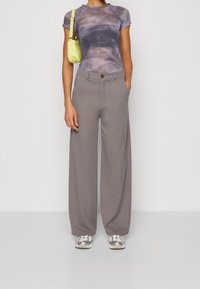 Grijze wijde broek met een knoop aan de voorkant, gecombineerd met een aansluitend tie-dye shirt in paarse tinten en een kleine gele schoudertas.