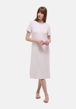 Femme debout pieds nus portant une robe midi simple, rose clair, à manches courtes, sur un fond blanc uni.