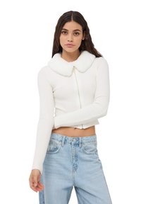 Biały sweter prążkowany z kołnierzem z sztucznego futra, zapinany na zamek z przodu, z długimi rękawami, w połączeniu z jasnoniebieskimi dżinsami z wysokim stanem.