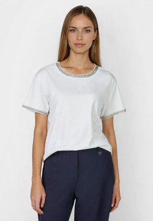 Frau trägt ein weißes kurzärmeliges Oberteil mit silberner Perlenverzierung und eine marineblaue Hose mit kleinem Herzanhänger an der Taille.