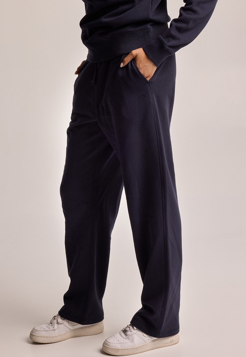 Námořnické modré tepláky vyrobené z měkké tkaniny, s bočními kapsami a uvolněným střihem. V páru s bílými sneakerami.