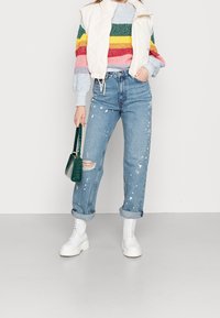 Gilet matelassé blanc sur un pull rayé multicolore, jeans déchirés bleu clair avec éclaboussures de peinture, et bottes blanches épaisses. Sac à main vert.