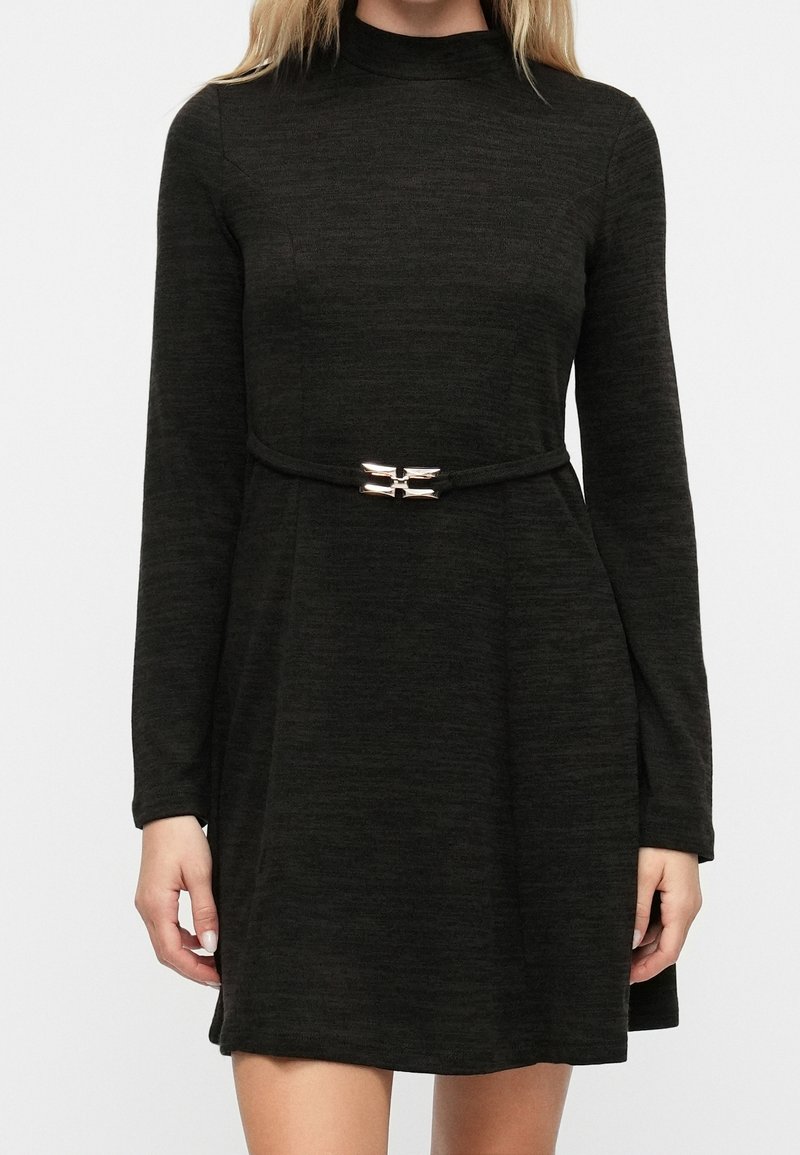 Femme portant une robe à manches longues, gris foncé, à col montant avec une fine ceinture noire ornée d'une boucle argentée à la taille.