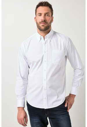 Mann mit kurzem Haar und Bart, der ein weißes langärmeliges Hemd mit Knöpfen und dunkle Jeans trägt, steht vor einem schlichten Hintergrund.