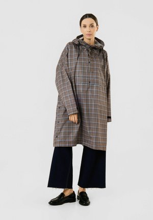 Femme portant un manteau ample à carreaux marron avec une capuche, un pantalon noir large et court, et des mocassins noirs, debout devant un fond uni.