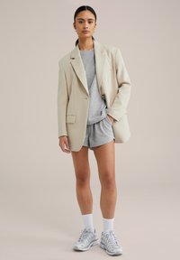 Beige oversized blézer, szürke póló és sort. A modellen fehér zoknik és világosszürke sportcipők vannak. Sima textúra, szabott dizájn.