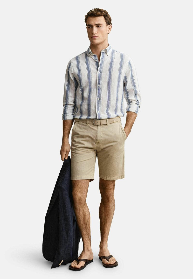 Mann trägt ein blau-weiß vertikal gestreiftes Hemd mit Knöpfen, beige Shorts mit Gürtel, hält eine dunkle Jacke in einer Hand und hat Flip-Flops an den Füßen.