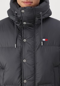Mørk grå dunjakke med høj krave, sølv hardware, frontknapper og et lille logopatch med røde og blå accenter.