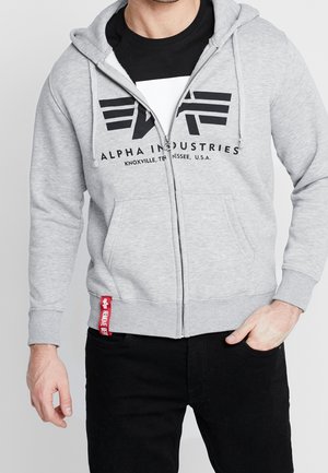 Mężczyzna noszący jasnoszarą bluzę Alpha Industries z zamkiem błyskawicznym na czarnej koszulce, z lewą ręką w kieszeni i prawym ramieniem w luźnej pozycji.