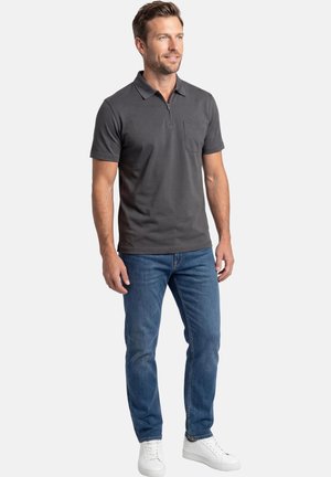 Mies seisoo tummanharmaa lyhythihainen polopaita, jossa vetoketjullinen kaulus, siniset farkut ja valkoiset tennarit yllään, yksiväristä taustaa vasten.