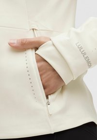 Light beige zip-up jacket featuring soft, smooth fabric, a zippered pocket, and reflective details on the sleeve with "LULULEMON" branding.