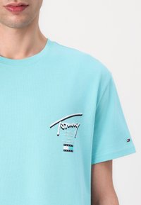 Tirkizna majica s kratkim rukavima od pamuka, s istaknutim bijelim grafičkim logotipom na lijevoj strani prsnog koša i suptilnim oznakama branda na rukavu.