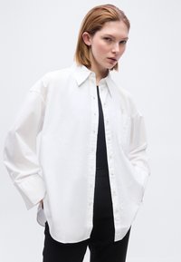 Νέα γυναίκα με κοντά μαλλιά, που φοράει ένα φαρδύ λευκό πουκάμισο με κουμπιά πάνω από μαύρο τοπ και μαύρο παντελόνι, με τα χέρια στις τσέπες, ουδέτερο φόντο.