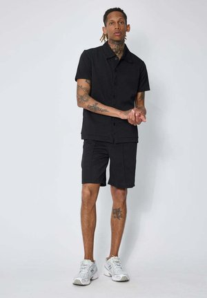 Homme avec des tatouages portant une chemise noire à manches courtes boutonnée, un short noir et des baskets blanches, debout devant un fond clair uni.