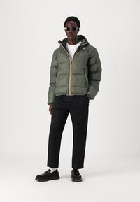 Giacca trapuntata verde con zip frontale, strisce gialle e nere, polsini elasticizzati e cappuccio. Abbinata a pantaloni e scarpe neri.