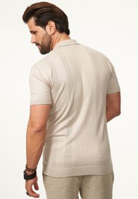 Beige Poloshirt mit kurzen Ärmeln, strukturiertem Muster und dezenten vertikalen Streifen. Hergestellt aus leichtem Strickmaterial.