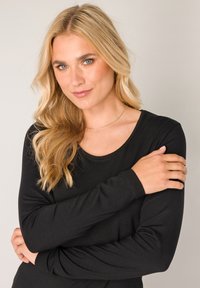 Zwart shirt met lange mouwen en een ronde halslijn. Zachte stof met een gladde textuur. Model met gekruiste armen, waardoor de mouwlengte en pasvorm zichtbaar zijn.