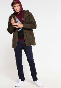 Maslinasto zelena jakna s kapuljačom preko bordo-sive hoodice. Tamnoplave hlače i bijele tenisice. Jednostavan dizajn s minimalnim hardverom.