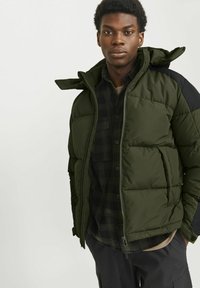 Olivengrønt puffer-jakke med sorte detaljer, quiltet design, lynlås foran, aftagelig hætte og frontlommer over en ternet skjorte.
