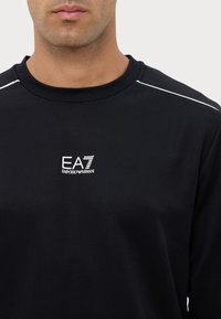 Maglietta nera a maniche lunghe realizzata in tessuto liscio con scollo rotondo. Presenta un logo EA7 bianco e sottili accenti bianchi sulle spalle.