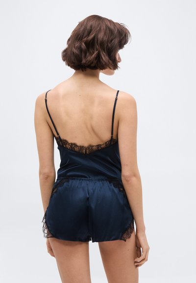 Žena s krátkymi hnedými vlasmi v námornícko modrom saténovom komplete tielko a šortky s čiernou čipkovou ozdobou, pohľad zozadu.