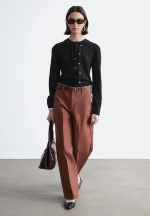 Kvinde iført sort knappet cardigan, brune højtaljede bukser, sorte flade sko, solbriller, ringøreringe, bærende på mørkebrun håndtaske, gående på gråt gulv.