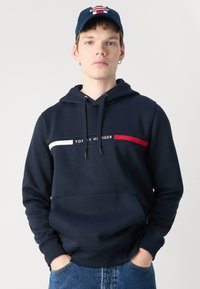 Tamsiai mėlyna džemperis su kišenę priekyje, su balta ir raudona horizontalia juostele ir "TOMMY HILFIGER" logotipu per krūtinę. Medžiaga – medvilnės mišinys.