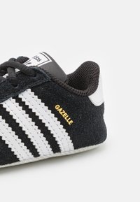 Černé semišové tenisky s bílými pruhy, texturovaným černým síťovaným jazykem a zlatým nápisem "Gazelle". Disponují bílou gumovou podrážkou.