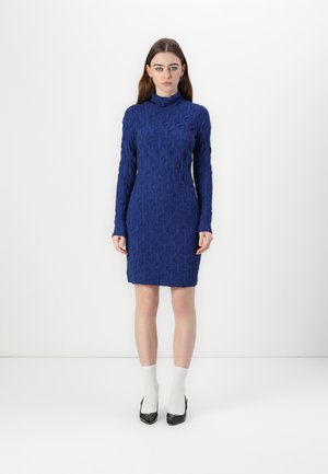 Robe en tricot bleu avec un col haut, des manches longues, un motif géométrique texturé, une silhouette ajustée et une longueur au genou. Talons hauts noirs.