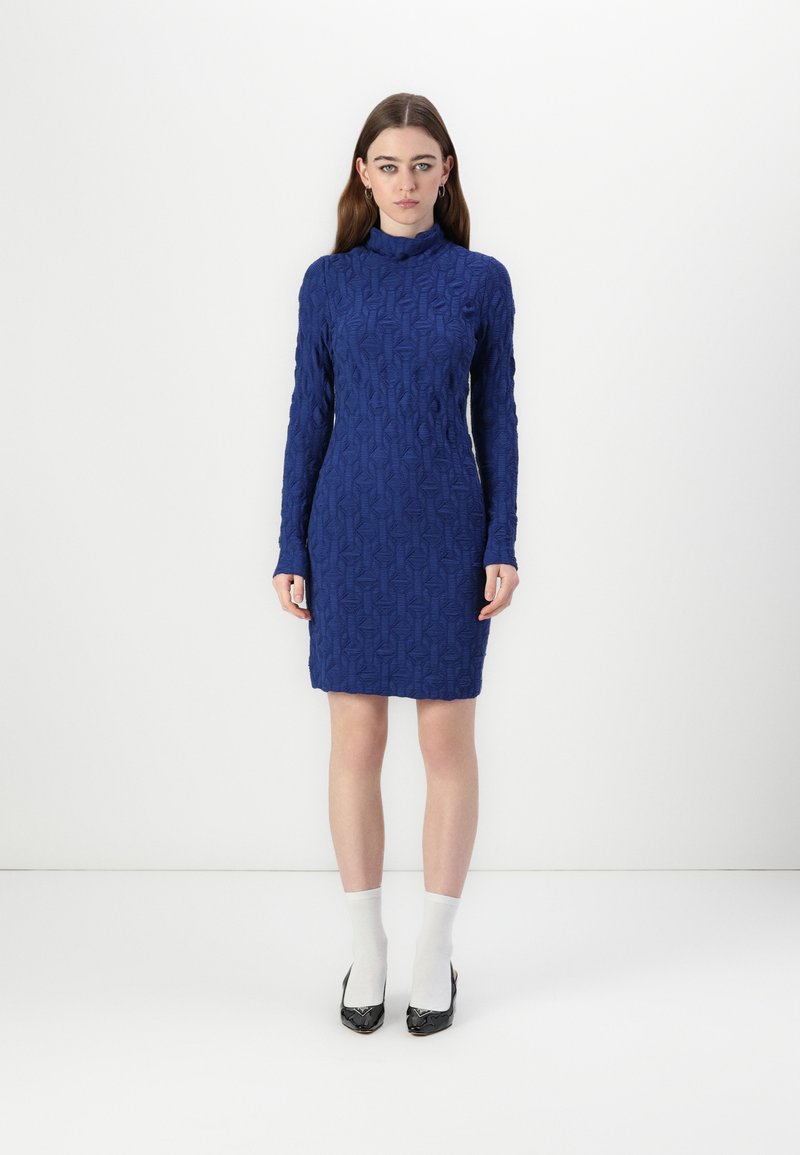 Rochie tricotată albastră cu guler înalt, mâneci lungi, model geometric texturat, siluetă ajustată și lungime până la genunchi. Pantofi cu toc înalt negri.