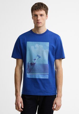 Młody mężczyzna w niebieskim t-shircie z grafiką ręki trzymającej małą szklankę na tle nieba i morza.