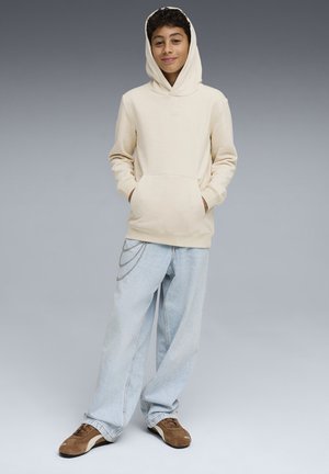 Chłopiec stoi z rękami w kieszeniach, ubrany w beżową bluzę z kapturem, jasnoniebieskie szerokie dżinsy z łańcuchami oraz brązowe sneakersy na szarym tle.