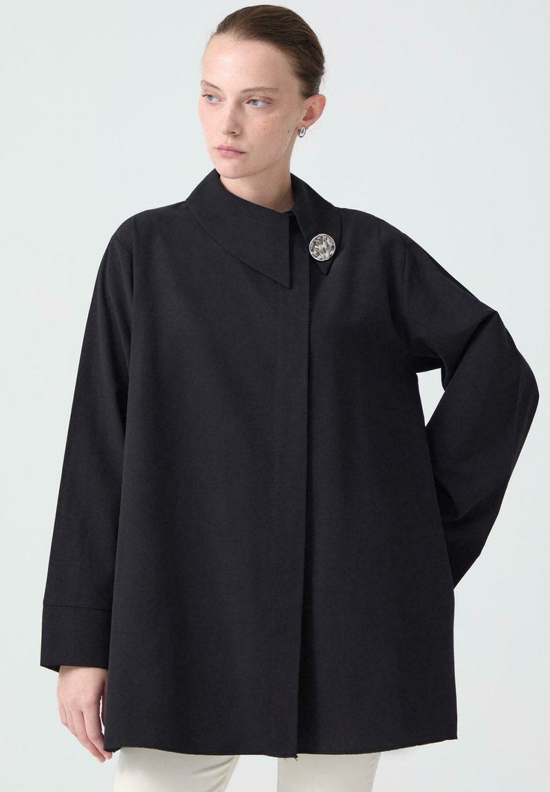 Black collared shirt with long sleeves, made from a smooth fabric, featuring a decorative brooch and a straight cut at the hem.