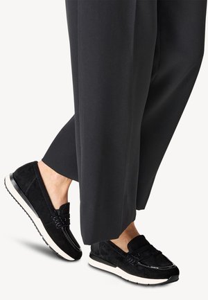 Scarpe slip-on in suede nera con suola bianca, caratterizzate da una punta liscia, dettagli di cucitura e un design a profilo basso. Indossate con pantaloni neri larghi.