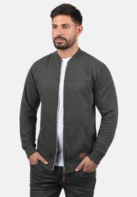 Mørkegrå zip-up sweatshirt med krageløs design, forrettede lommer og et læderlogo. Båret over en hvid t-shirt.
