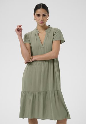 Sötét hajú nő világoszöld, rövid ujjú, réteges ruhában, arany karikafülbevalóval és arany karkötővel, egyszínű háttér előtt áll.