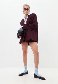 Blazer oversize bordeaux sur une chemise bleu clair, associé à des ballerines noires et des chaussettes bleu clair. Sac à main noir et lunettes de soleil rondes violettes.
