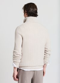 Pull en tricot beige avec un col montant, texture côtelée et manches longues. Détails notables à l'ourlet et aux poignets avec une chemise blanche en dessous.