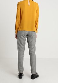 Senapsgul blus med en rund nyckelhålsdetalj på baksidan, i kombination med grå rutiga byxor. Byxorna har en skräddarsydd passform och en subtil lyster.