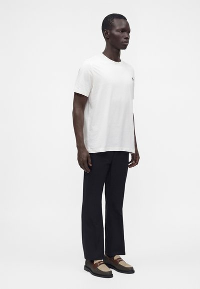 Homme debout portant un t-shirt blanc uni à manches courtes, un pantalon noir droit, et des mocassins bicolores marron et beige.