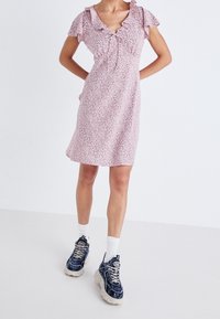 Rosa blommig klänning med volangärmar, v-ringning och samlad midja. Bärs med mörka sneakers och vita strumpor för kontrast.