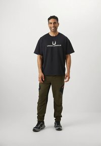 Črna bombažna majica s tiskanim belim logotipom "Under Armour", združena z olivno zelenimi trenirkami z črnimi poudarki in rdečimi detajli.