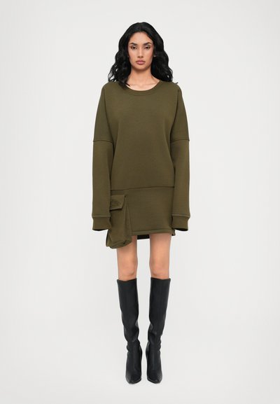 Robe oversize vert olive à manches longues, en tissu texturé, avec un détail de poche sur le côté. Associée à des bottes noires montantes.
