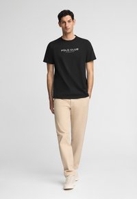 Svart bomullströja med "POLO CLUB"-logotyp i vitt, tillsammans med beige byxor och vita sneakers, presenterad mot en neutral bakgrund.