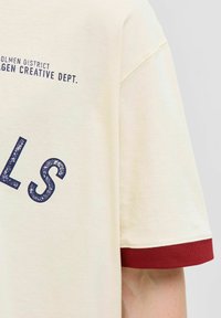 Bēša īsu piedurkņu krekls ar tumši sarkanu piedurkņu apdari un tumši zilu tekstu, kas lasāms "HOLMEN DISTRICT COPENHAGEN CREATIVE DEPT."