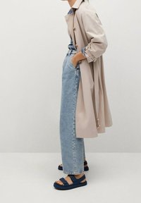 Beige trenchcoat, ontspannen lichtblauwe denim jeans en marineblauwe sandalen met klittenbandsluitingen. Glad stof en losvallend ontwerp.