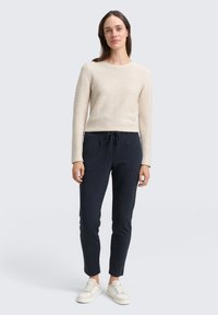 Beige ribbade långärmade topp i kombination med marinblå dra-mudden-byxor. Modellen har på sig ljusa sneakers. Enkel, figursydd design, avslappnad look.