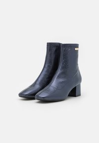 Botines azul marino con un acabado brillante, punta cuadrada y tacón grueso. Presentan una cremallera lateral para facilitar su uso.