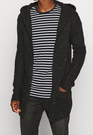 Homme portant un cardigan noir à capuche en maille par-dessus une chemise rayée noir et blanc avec un jean noir, debout devant un fond uni.