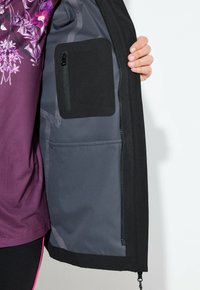 Sort jakke med en grå indvendig foring, der har en lynlåslomme og en større åben lomme. Stoffet ser glat og holdbart ud.